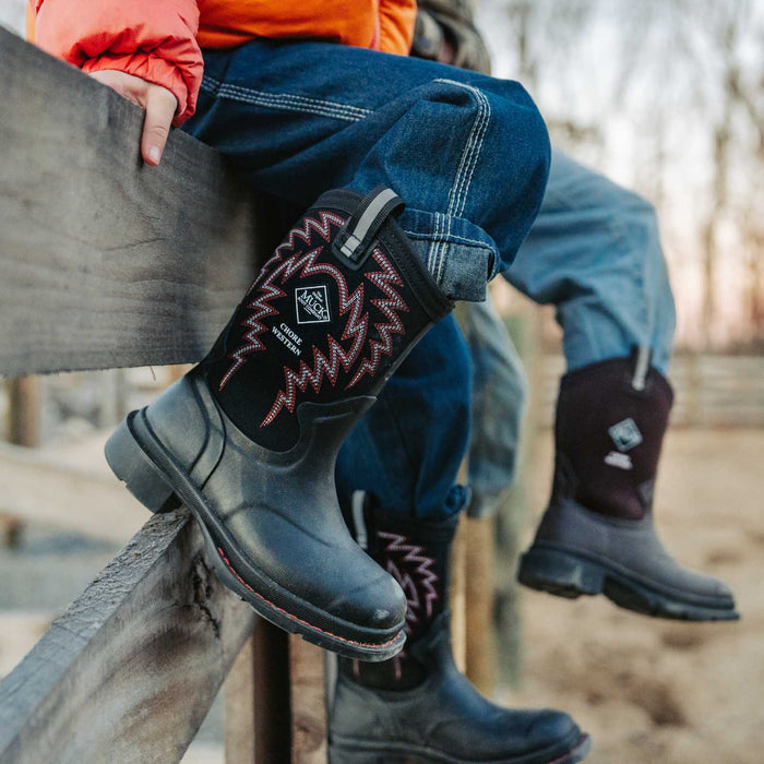 Person wearing black boots with red patterns, sitting on a wooden fence outdoors.