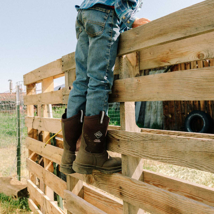 Person wearing jeans and boots standing on a wooden fence with a rural background
