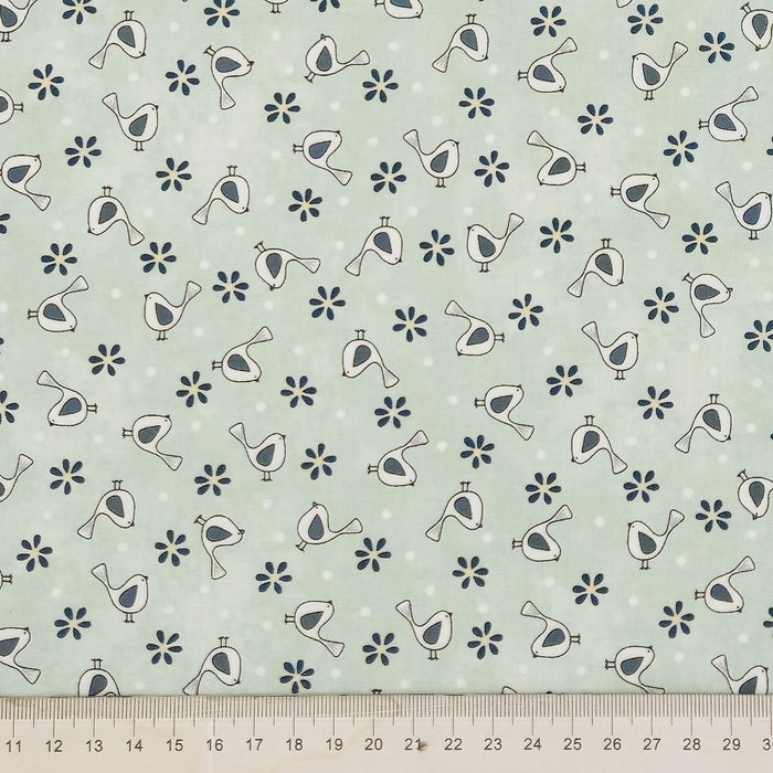 A close-up of a fabric pattern featuring small white birds and dark blue flowers on a pale sage green background, with a ruler along the bottom edge showing scale.