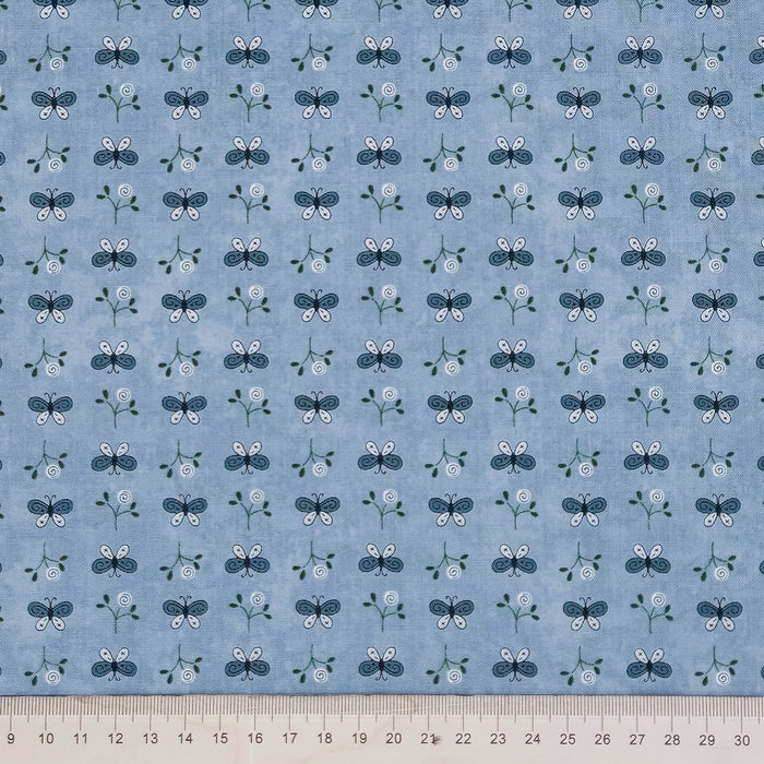 Light blue fabric with a repeating pattern of small blue, white, and green floral motifs, shown flat with a measuring ruler along the bottom edge.