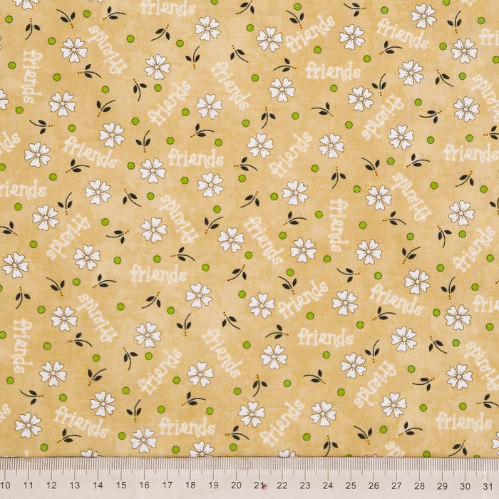 A close-up of a fabric pattern featuring white daisy flowers, green dots, and the word 'friends' repeated diagonally in gray, on a warm tan cotton quilting fabric. A silver metal ruler is visible along the bottom edge to indicate scale.