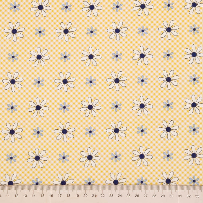 Yellow and white gingham fabric with a repeating pattern of white daisies with dark blue centers and small light blue flowers, shown with a ruler at the bottom to indicate scale.