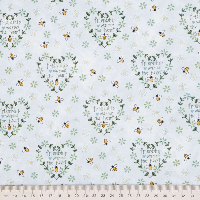 A close-up of patterned fabric with a repeating design of small yellow-and-black bees, green leafy wreaths, and tiny white flowers on a pale background. The center of several motifs reads 'friendship warms the heart.