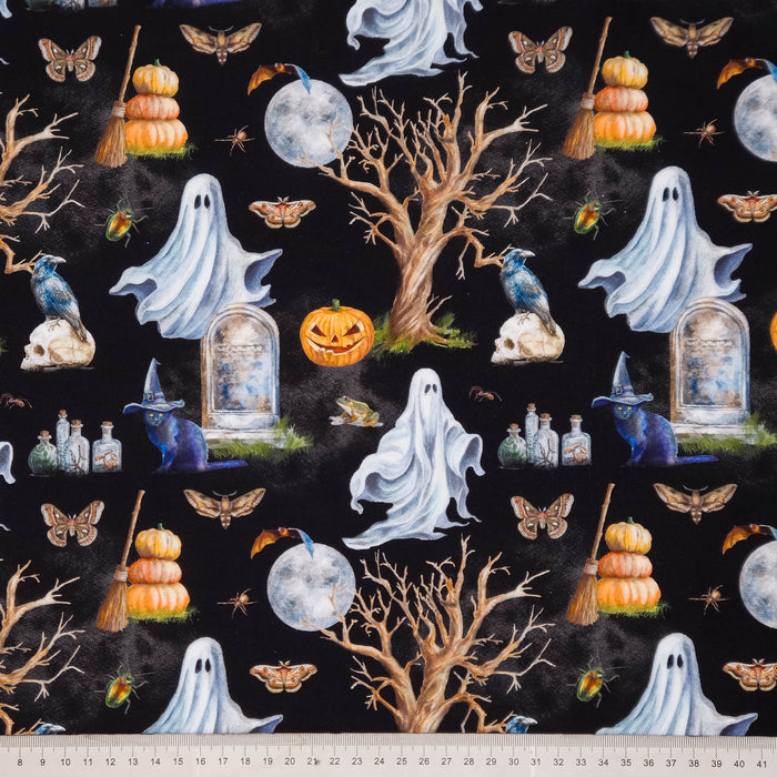 Halloween-themed fabric print featuring repeating motifs of white ghosts, orange pumpkins, bare trees, tombstones, bats, skulls, cauldrons, and a black background, with a ruler along the bottom edge showing scale.