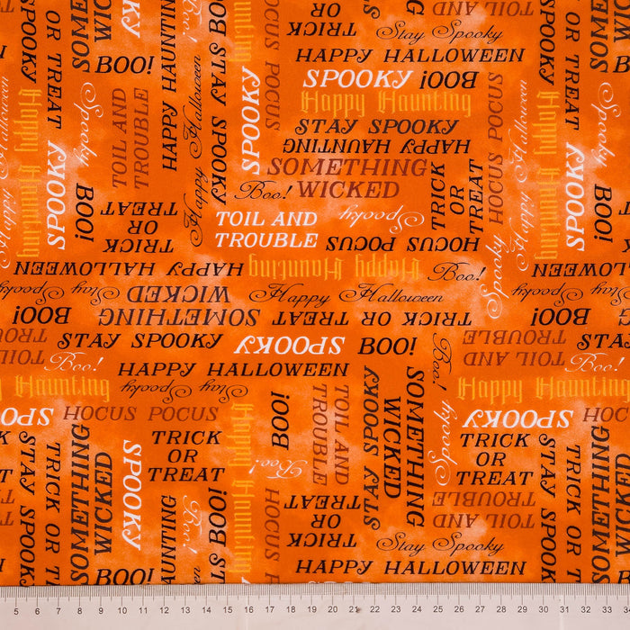Bright orange Halloween-themed fabric with a repeating pattern of words and phrases like 'BOO', 'SPOOKY', 'HAPPY HALLOWEEN', 'TOIL AND TROUBLE', and 'WICKED' in black and white text, shown with a white measurement ruler along the bottom edge.