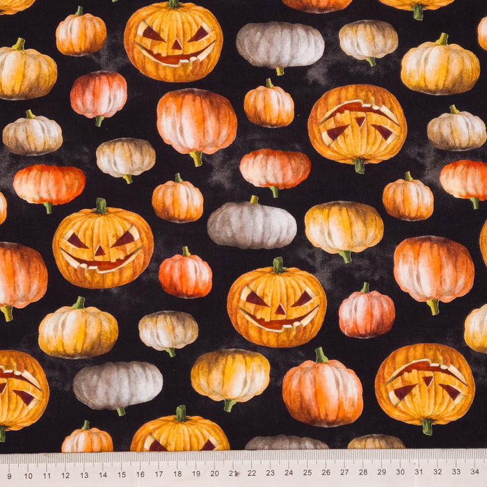 Pumpkin patterned fabric with illustrated jack-o'-lanterns and small pumpkins in orange, peach, cream, and gray on a black background, with a measuring ruler along the bottom edge.