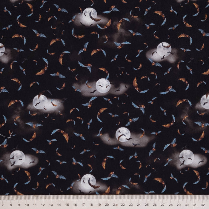 Black fabric with a repeating pattern of flying bats and moon phases in blue, orange, and white, with a white ruler visible along the bottom edge.