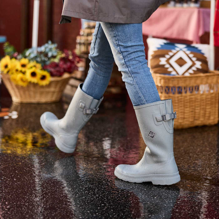 Person wearing tall gray women's adjustable rain boots on a wet pavement with a market background