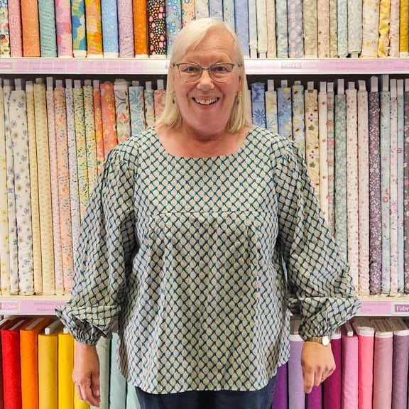 Person wearing a patterned shirt and blue pants standing in front of fabric shelves.