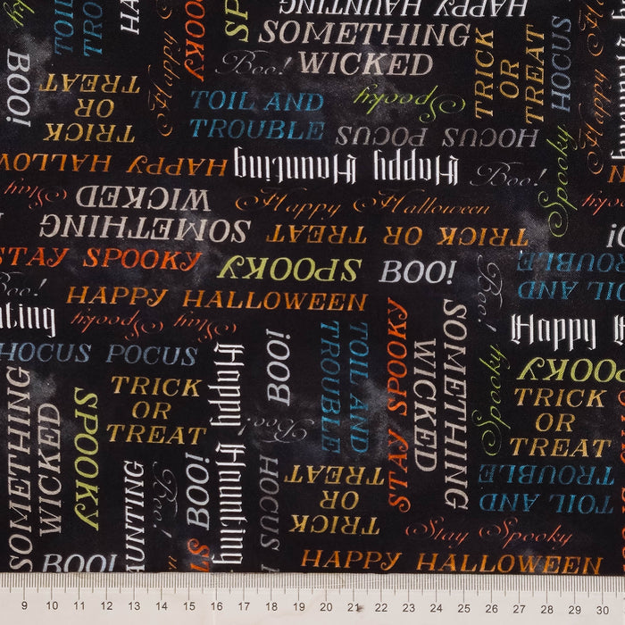 A close-up of Halloween-themed fabric with a black background and scattered white, orange, blue, and yellow text words like 'BOO!', 'HAPPY HALLOWEEN', 'WICKED', and 'SPOOKY'. A metal ruler is visible along the bottom edge.